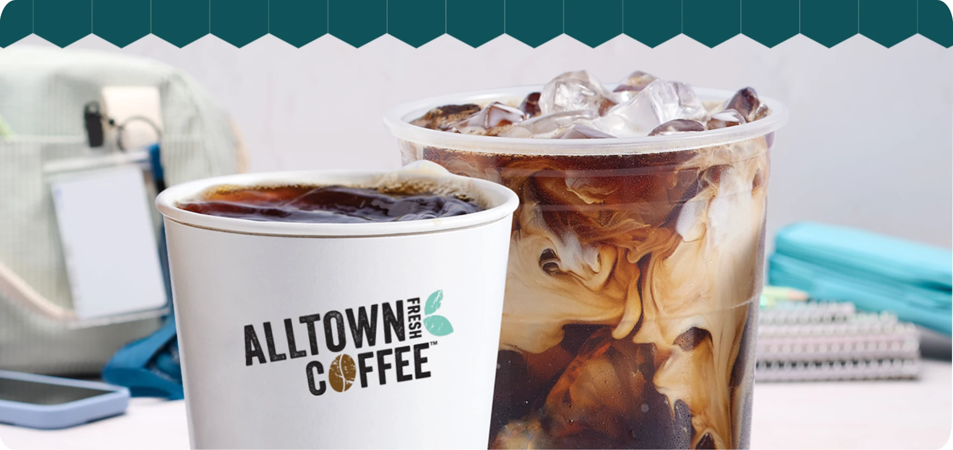 Two coffee drinks on a desk: a white Alltown Coffee cup and a large iced coffee with caramel swirls in a clear cup.